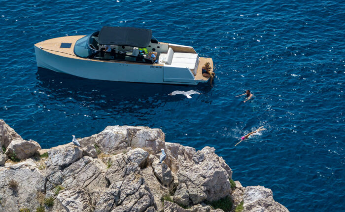 A boat is anchored near a rocky coastline in clear blue water during one of the idyllic private boat tours in Dubrovnik. Two people are swimming beside the boat while seagulls sit on the rocks.