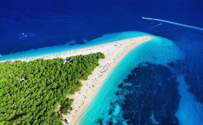 Aerial view of a narrow sandy beach stretching into the sea with dense green foliage on one side and clear blue water on the other. A speedboat, possibly part of Dubrovnik's private boat tours, creates a wake in the dark blue sea.