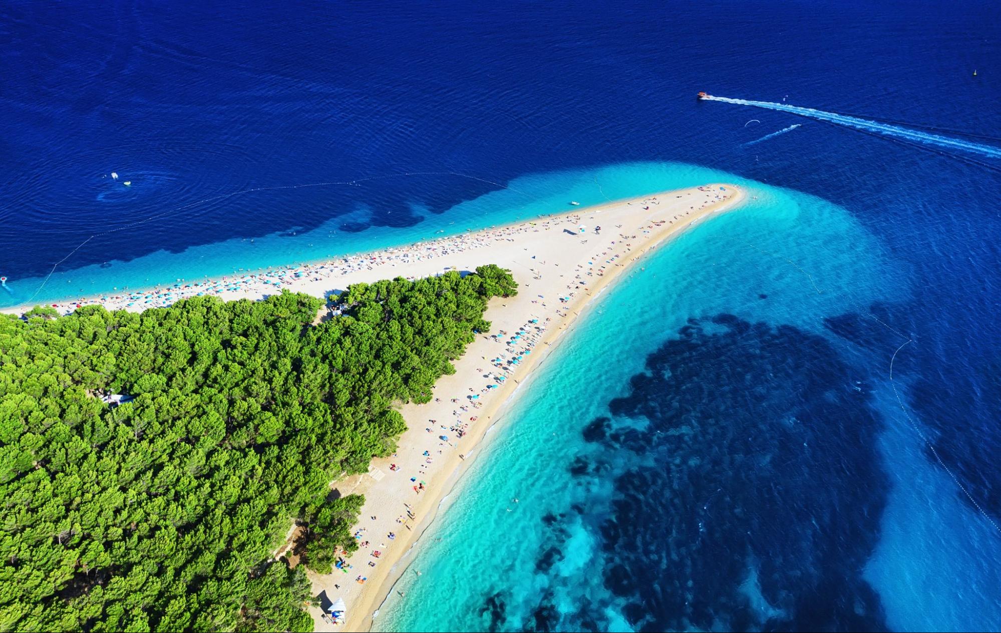 Aerial view of a narrow sandy beach stretching into the sea with dense green foliage on one side and clear blue water on the other. A speedboat, possibly part of Dubrovnik's private boat tours, creates a wake in the dark blue sea.