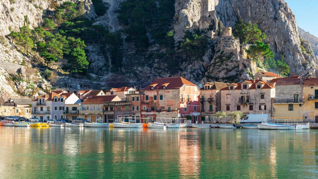 A quaint coastal village with colorful houses and boats docked on calm water, backed by a rocky hillside with greenery.