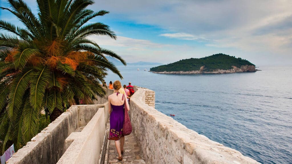 A woman in a purple dress walks along a stone pathway by the sea, with an island visible in the distance and a large palm tree nearby.