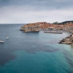 A coastal view of Dubrovnik, Croatia, showing historic walls and buildings, a few anchored boats, a small beach, and a calm sea under a cloudy sky.