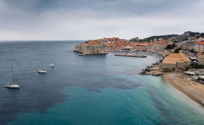 A coastal view of Dubrovnik, Croatia, showing historic walls and buildings, a few anchored boats, a small beach, and a calm sea under a cloudy sky.