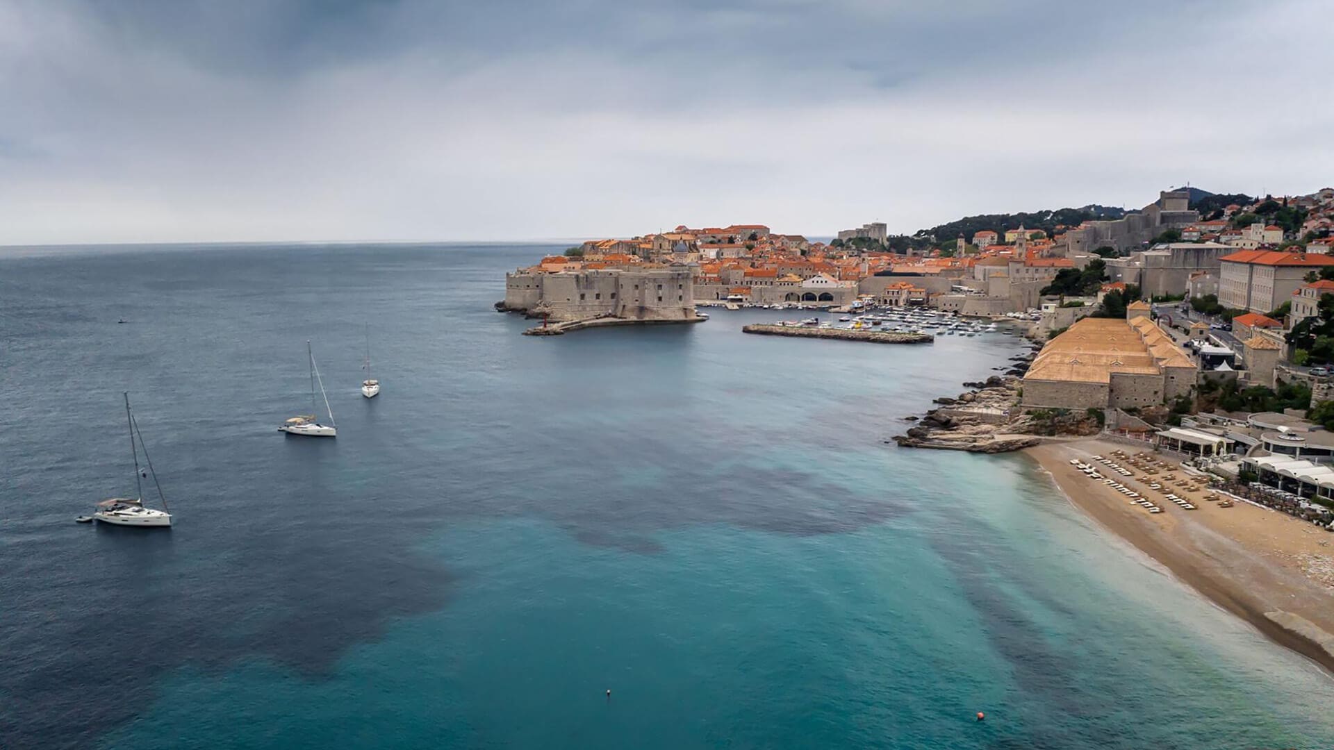 A coastal view of Dubrovnik, Croatia, showing historic walls and buildings, a few anchored boats, a small beach, and a calm sea under a cloudy sky.