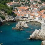 A coastal town with red-roofed buildings, surrounded by clear blue water, rocky cliffs, and kayakers in the sea.