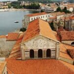 Aerial view of a coastal town in Croatia with red-tiled roofs, historic buildings, and the sparkling Adriatic Sea in the background.