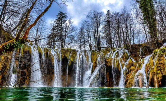 Numerous small waterfalls cascade over rocks into a clear green pool, surrounded by leafless trees on a partly cloudy day.