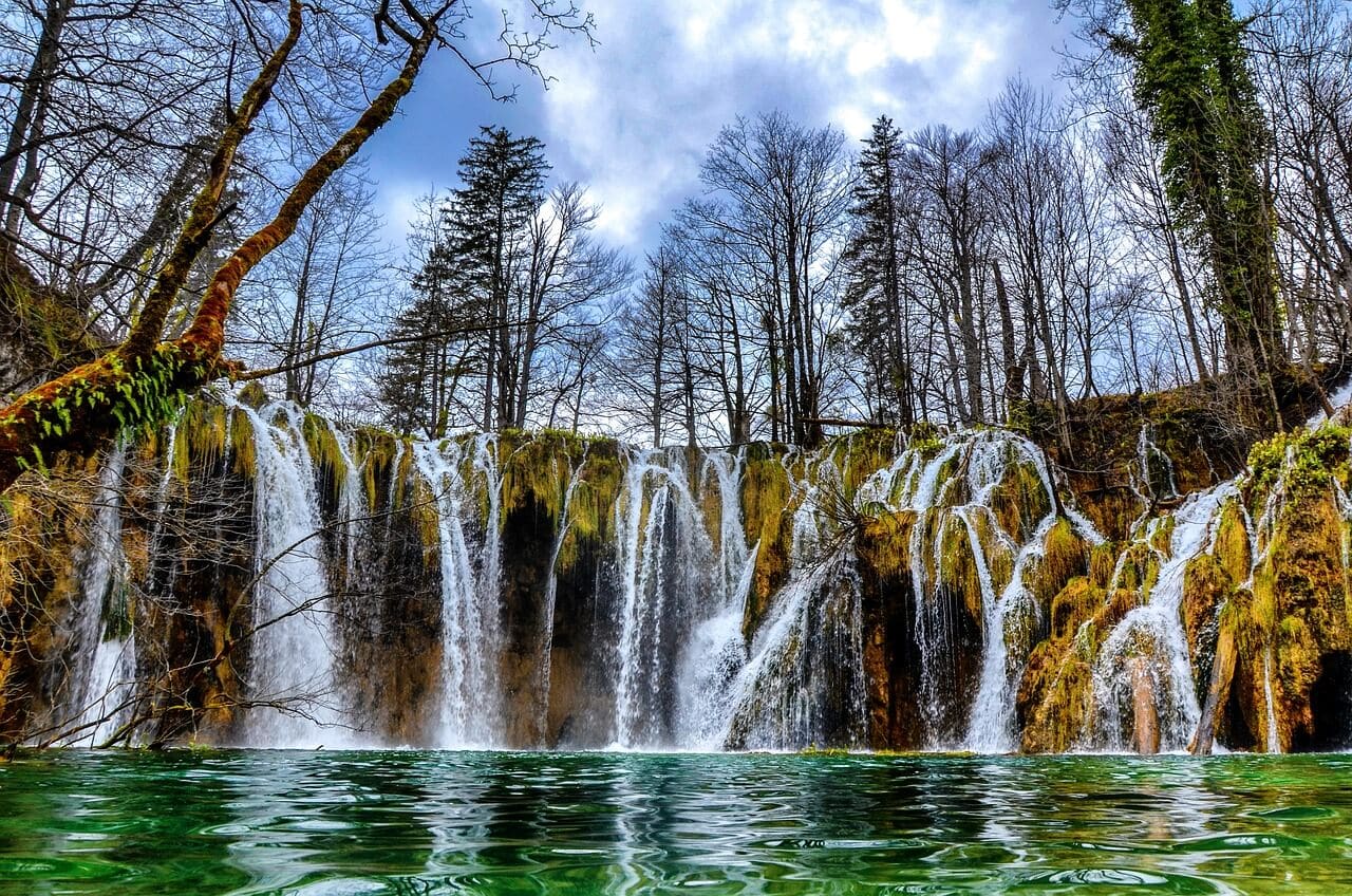 Numerous small waterfalls cascade over rocks into a clear green pool, surrounded by leafless trees on a partly cloudy day.