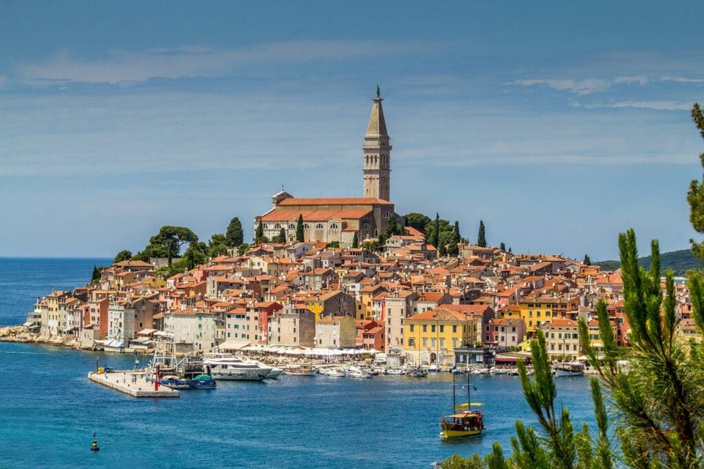 A coastal town in Croatia with a prominent bell tower surrounded by red-roofed buildings and docked boats, set against a blue sky and sea, reminiscent of the charm found in Dubrovnik's picturesque streets.