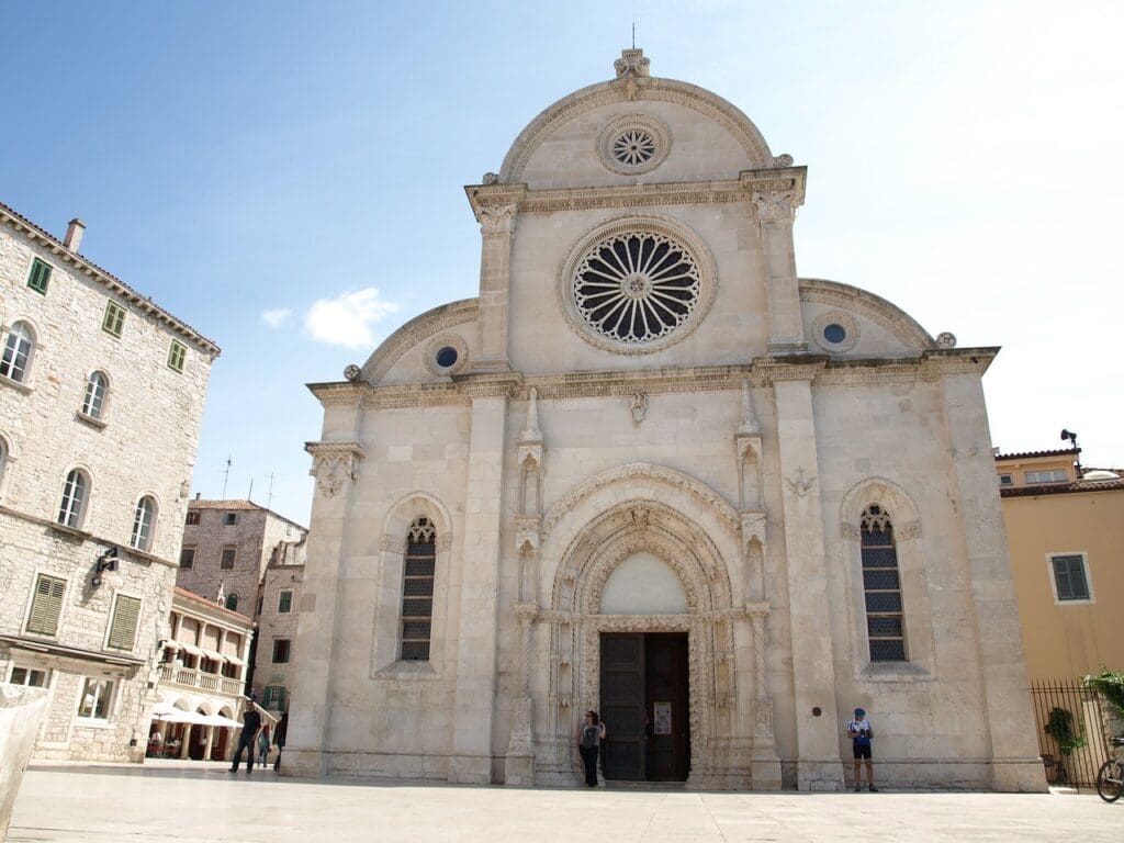 A historic stone cathedral with a large circular window stands under a clear sky in Dubrovnik, Croatia. People are seen walking and standing around the building's entrance, enjoying the charm of one of the most picturesque coastal towns.