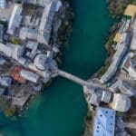 Aerial view of a bridge spanning a river, surrounded by historic stone buildings and streets. The greenish-blue water flows through the center of the town.