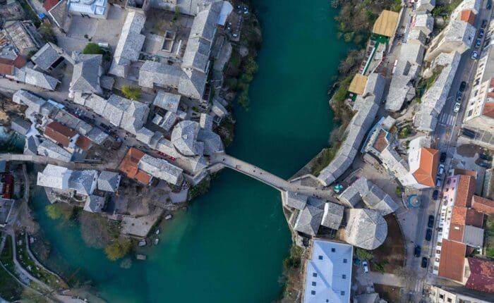 Aerial view of a bridge spanning a river, surrounded by historic stone buildings and streets. The greenish-blue water flows through the center of the town.