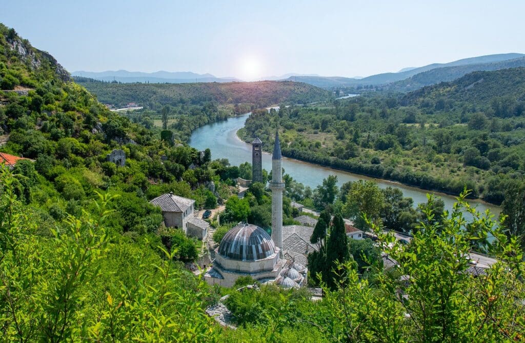 Aerial view of a river winding through a lush valley with hills. A prominent mosque with a dome and two minarets is in the foreground, surrounded by greenery.