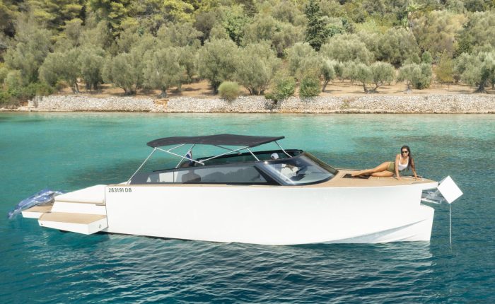 A woman in a swimsuit is lounging on the deck of a modern white motor yacht anchored in clear turquoise water near a forested shoreline.