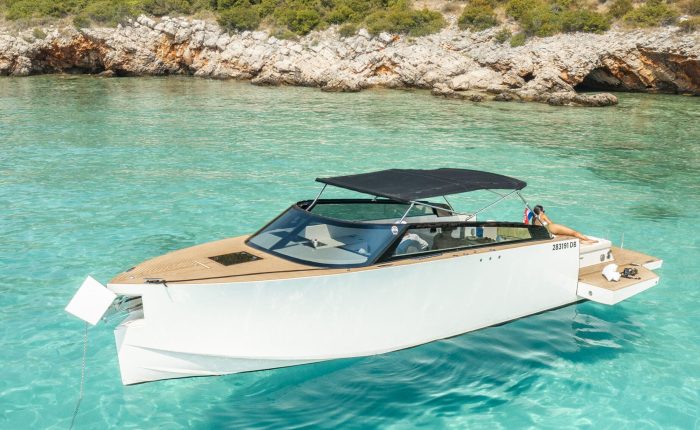 A modern white motorboat with a black canopy floats on clear turquoise water near a rocky, tree-lined shore.