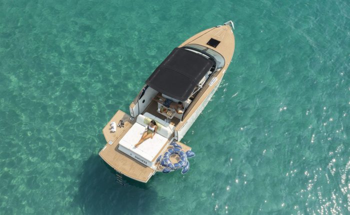 A person is sunbathing on the back of a small motorboat floating on clear turquoise water, with two inflatable rings nearby.