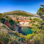 Scenic view of a coastal town with red-roofed buildings, surrounded by greenery and hills, beside clear blue water under a bright sky.