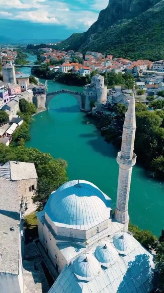 Aerial view of a historic bridge over a turquoise river, surrounded by buildings with red-tiled roofs, a minaret in the foreground, and lush green hills in the background.