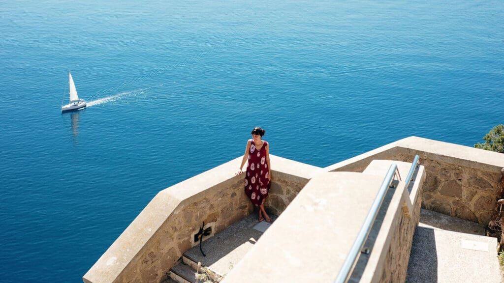 A person stands on a stone terrace overlooking the ocean with a sailboat in the distance.