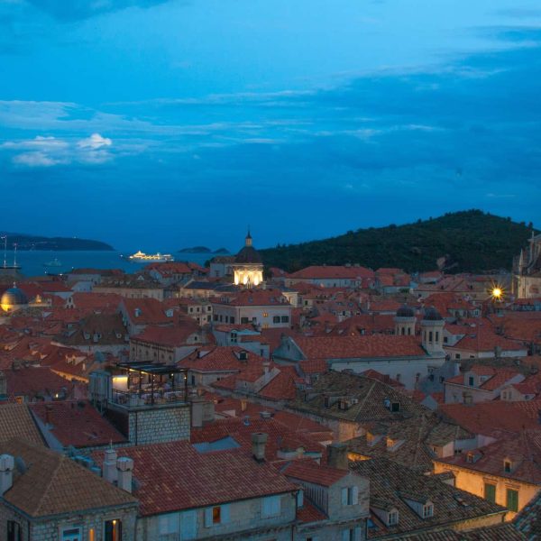 Evening view of a historic coastal city with glowing lights, red-tiled roofs, and a faintly lit church tower under a cloudy sky by the sea.