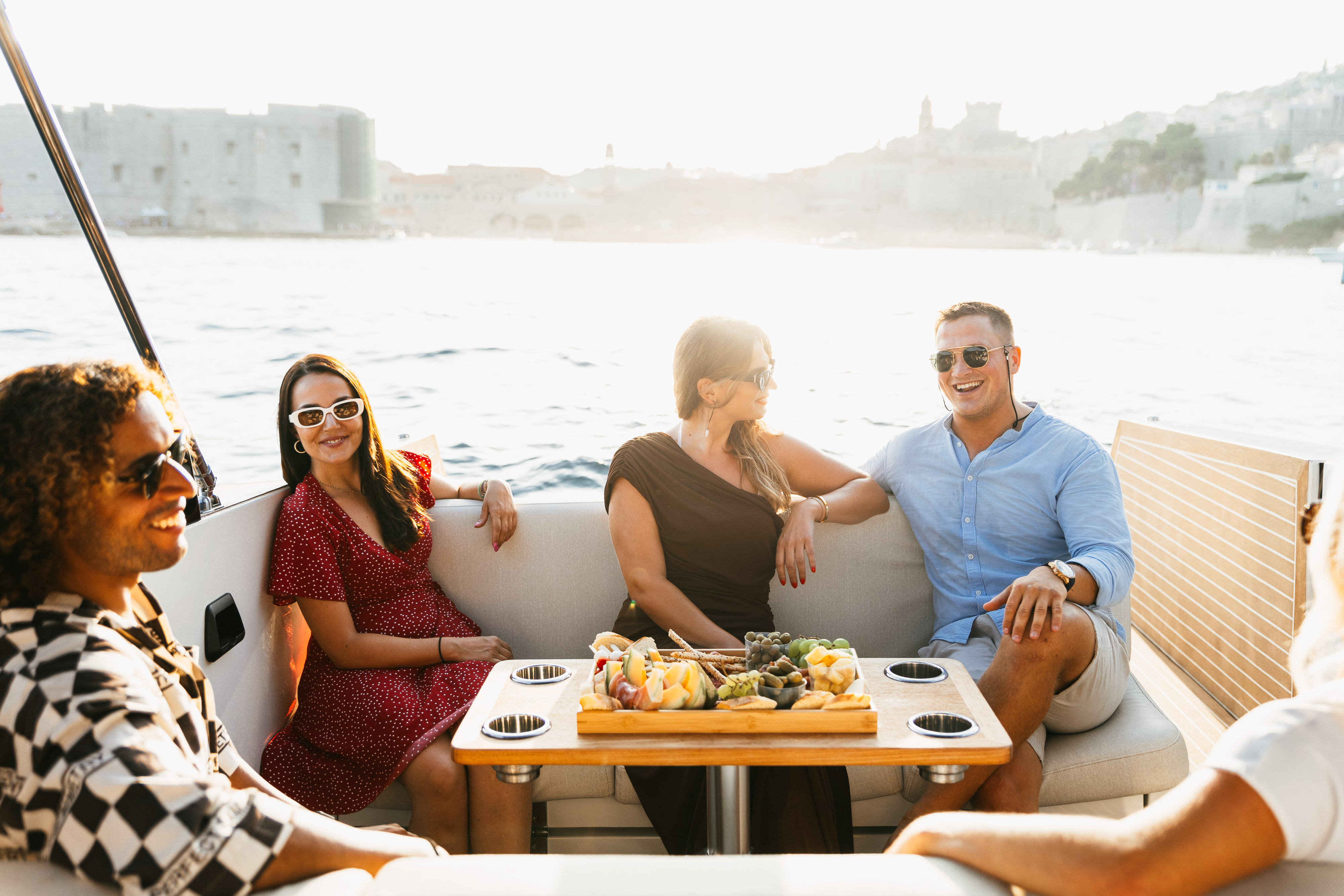 A group of four people is seated on a boat, savoring a platter of food against the backdrop of shimmering water and a stunning cityscape. As they laugh and chat, it seems there are no terms and conditions to their carefree enjoyment under the open sky.