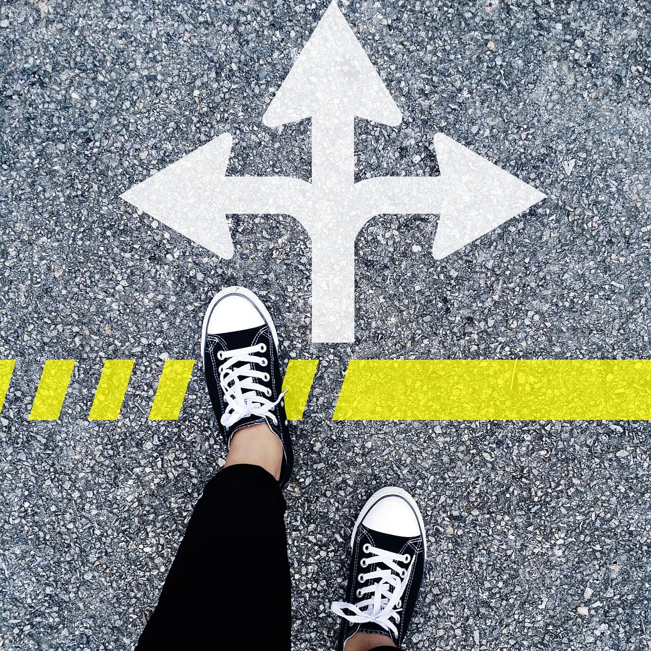 Feet in black sneakers stand on a road with a white multi-directional arrow and a yellow line, ready to journey onward—traveling life's many paths, much like exploring the charming streets of Croatia.