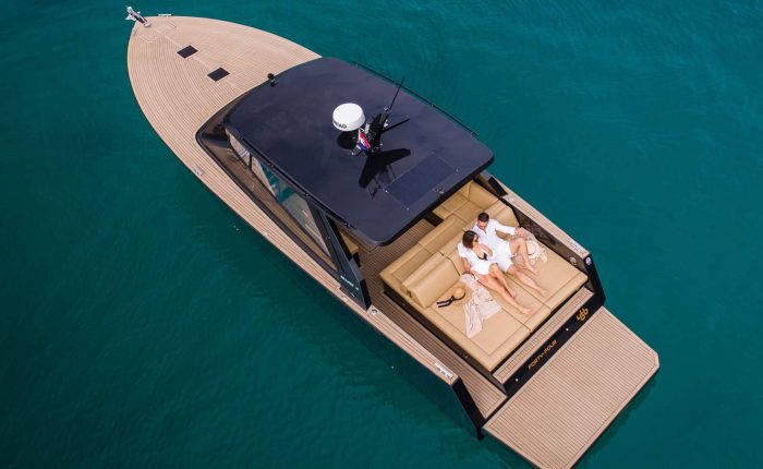 Aerial view of a couple relaxing on a luxury boat with beige decking, surrounded by clear turquoise water.