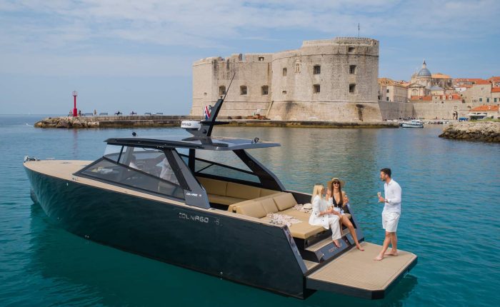 A modern black yacht with three people onboard is docked near a historic stone fortress and lighthouse on a sunny day.