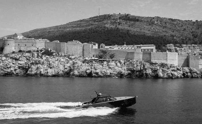 A motorboat moves across the water in front of a walled city built on rocky cliffs, with a large hill and trees in the background.