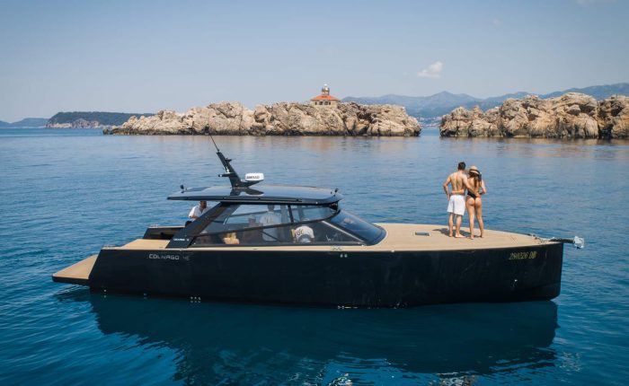A modern black yacht floats on calm blue water near rocky islands; two people stand at the bow looking at the scenery.