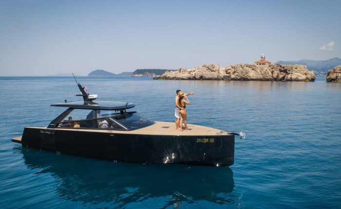 A couple stands on the deck of a sleek, modern yacht on clear blue water, pointing towards distant rocky shores with a small building.