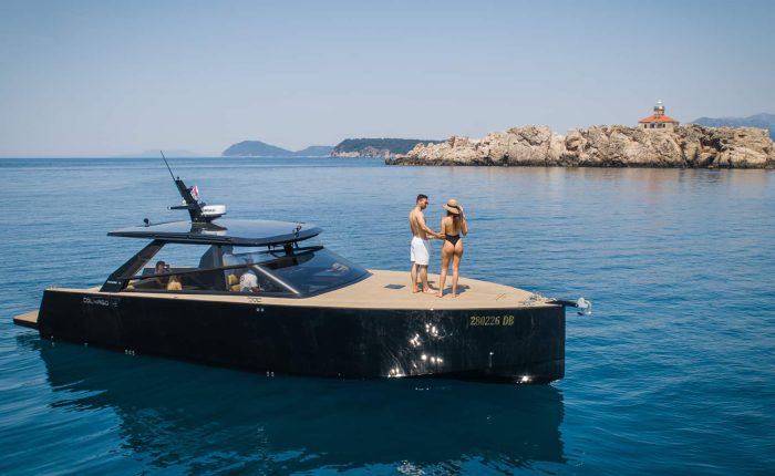 A modern black yacht with people on board floats on calm blue water near a rocky island with a small building in the background.