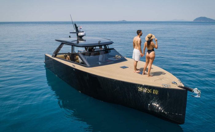 A man and a woman in swimwear stand on the deck of a modern black yacht floating on calm blue water.