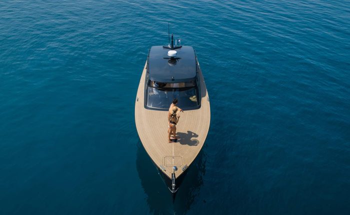 A person stands on the deck of a modern yacht floating on calm, deep blue water, viewed from above.
