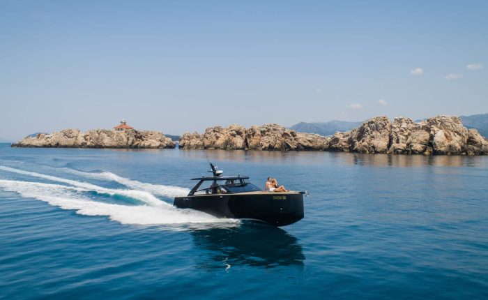 A black motorboat speeds across clear blue water with rocky islands and a lighthouse in the background.