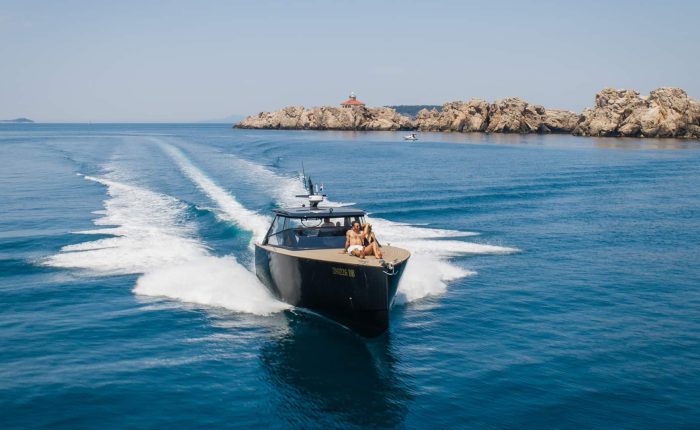 A motorboat travels across clear blue water, leaving a wake behind. Rocky coastline with small lighthouse in the background under a clear sky.