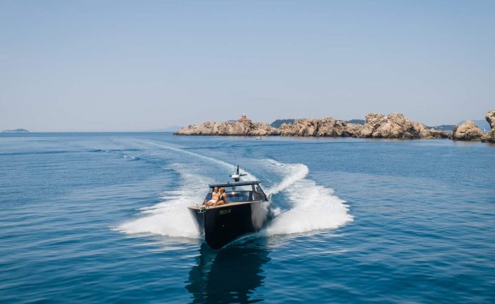 A black speedboat travels on clear blue water, creating a white wake behind it. Rocky islands and a lighthouse are visible in the background under a clear sky.