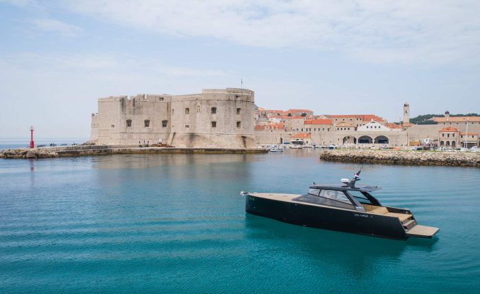 A modern black yacht floats in clear blue water near an old stone fort and historic buildings along the coastline under a partly cloudy sky.