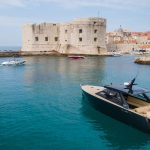 A modern motor yacht with people on board is anchored near the historic stone walls of Dubrovnik, Croatia, with other boats and the city in the background.