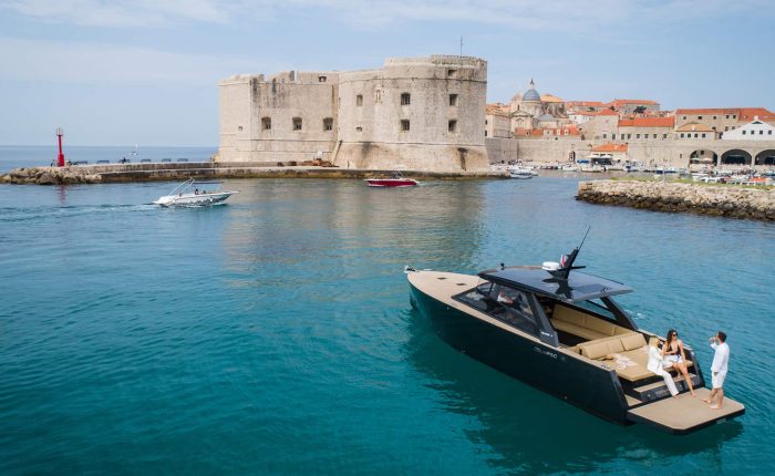 A modern motor yacht with people on board is anchored near the historic stone walls of Dubrovnik, Croatia, with other boats and the city in the background.