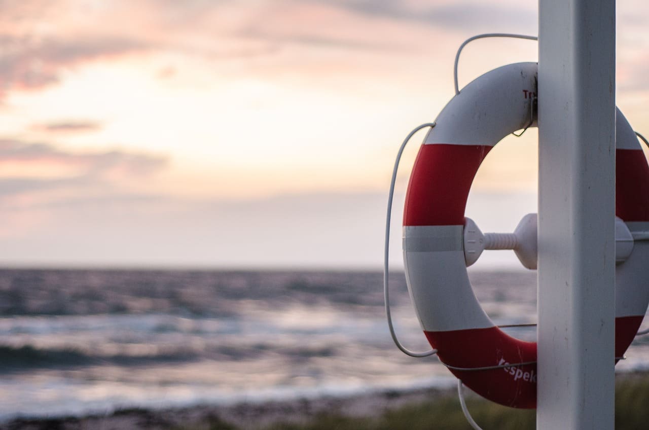 A red and white lifebuoy is mounted on a pole, overlooking a Croatian beach with waves under a cloudy sky at sunset, inviting those traveling to embrace the tranquil beauty.