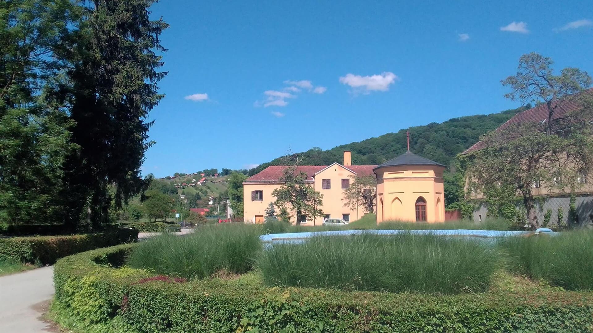 A sunny day with green hedges, grass, and a path in the foreground leads to yellow buildings with red roofs and hills in the background. Sparse clouds dot the clear blue sky, creating a perfect setting for wellness getaways near Croatia's natural springs.