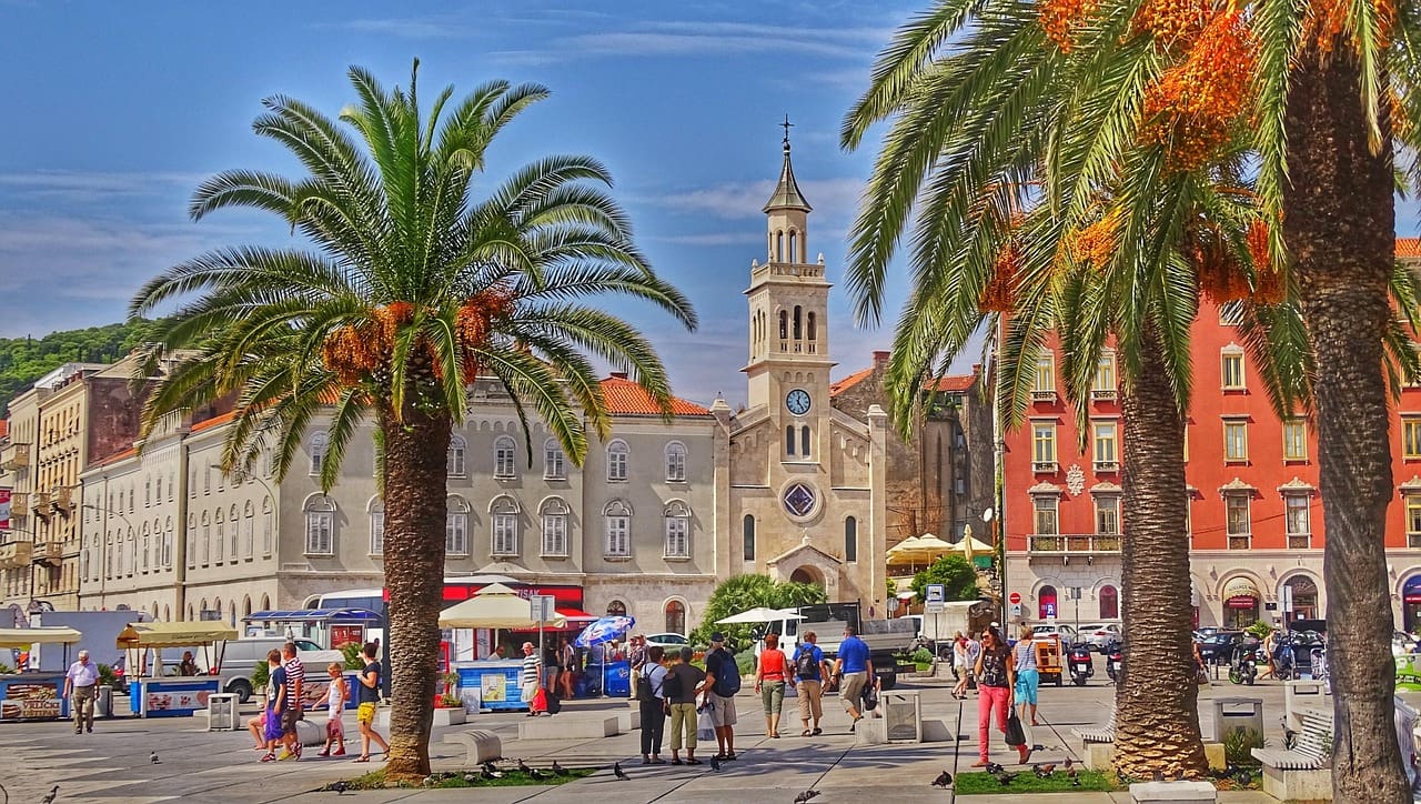 A sunny plaza with a church tower, surrounded by palm trees and colorful historic buildings, offers a picturesque spot for those traveling in Croatia. People walk and sit around the open, vibrant area, experiencing the unique charm of this lively destination.