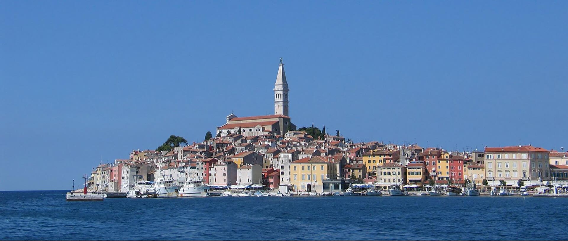 Coastal town with colorful buildings and a prominent church spire, set against a clear blue sky, viewed from the water—a perfect backdrop for wellness getaways featuring Croatia’s natural springs.