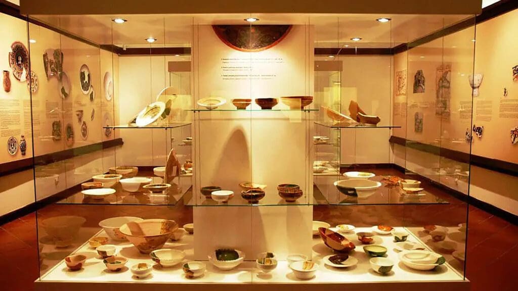 A well-lit museum display in Dubrovnik features three glass shelves showcasing various pottery artifacts and ceramic pieces. Ideal for day trips, the exhibit includes mounted information panels and additional pottery on the background walls.