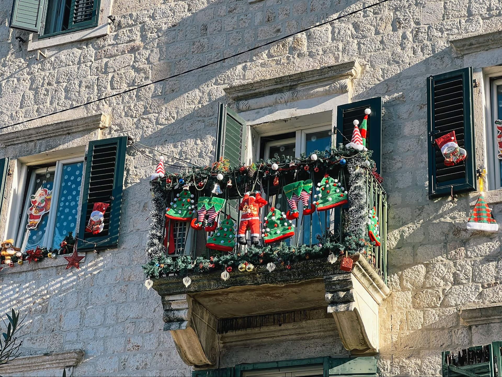 A stone building facade with green shutters in Dubrovnik displays festive holiday decorations on a balcony, including Santa figures, colorful garlands, and hanging ornaments against a sunny winter backdrop.