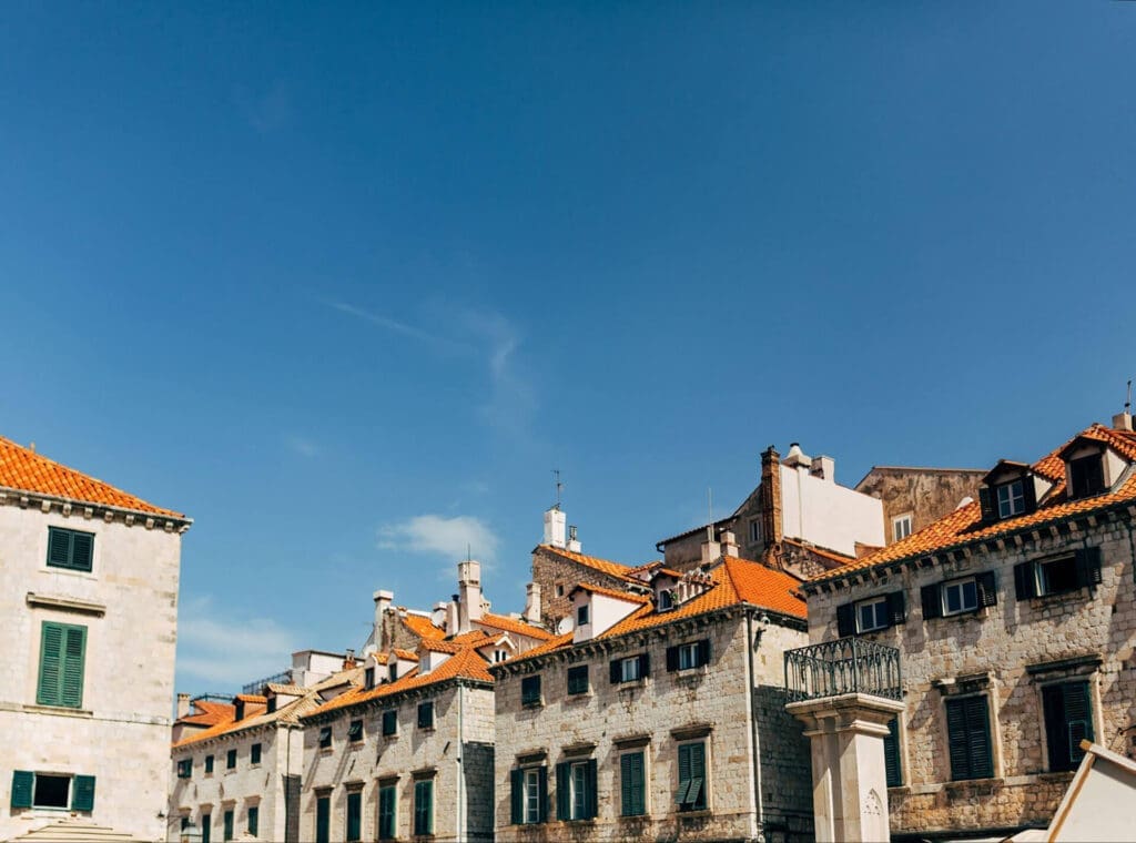 Historic stone buildings with red-tiled roofs under a clear blue sky. The architecture features small windows and a mixture of old and modern elements.
