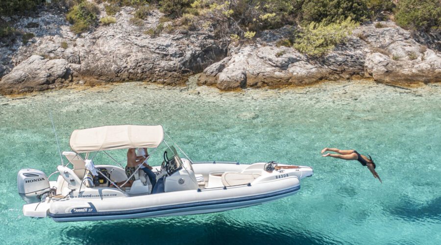 A person dives off a small white motorboat to swim in clear turquoise water near a rocky, tree-lined shore in Dubrovnik.