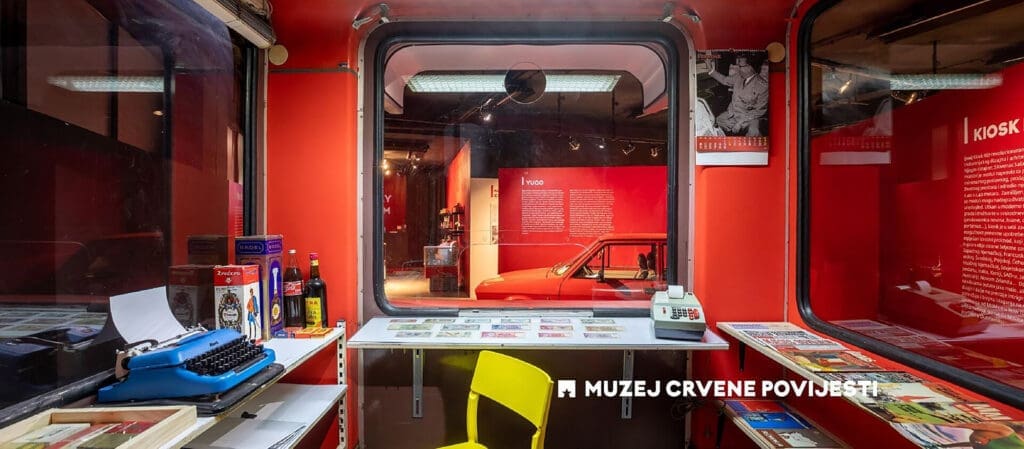 An exhibit at the Museum of Red History in Dubrovnik features a red room with a vintage typewriter, books, and postcards, alongside a yellow chair in the foreground. In the background, you'll find a red car and informational panels. Consider adding this unique museum to your day trips itinerary while exploring private boat tours around Dubrovnik.