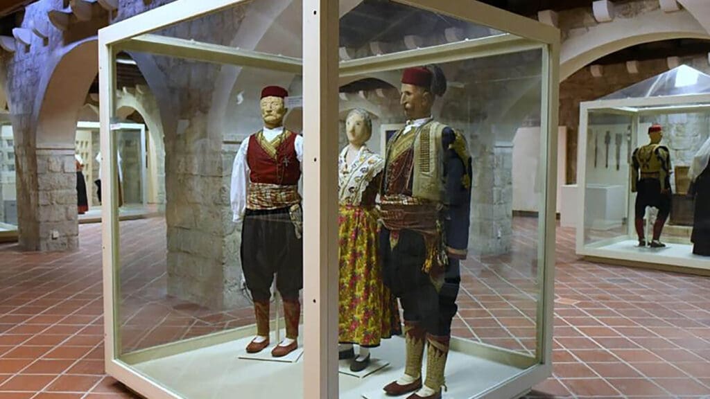 Three mannequins dressed in traditional attire are displayed in a glass case within a museum room featuring stone walls and tiled floors, offering a glimpse into Dubrovnik's rich cultural heritage.